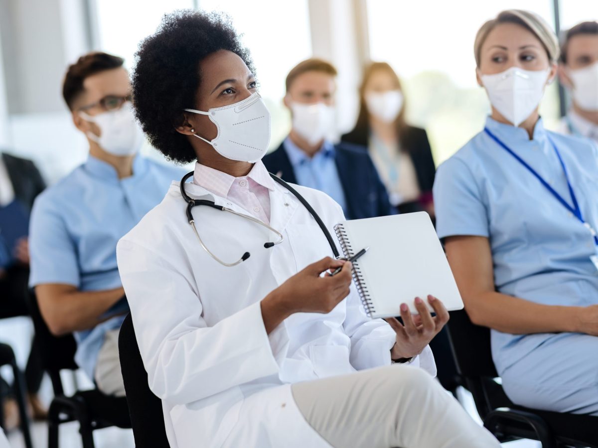 Happy black doctor pointing at her notebook while being on a seminar with group of colleagues during COVID-19 pandemic.