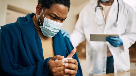 Worried black man with face mask sitting wrapped in blanket while doctor is visiting him at home.