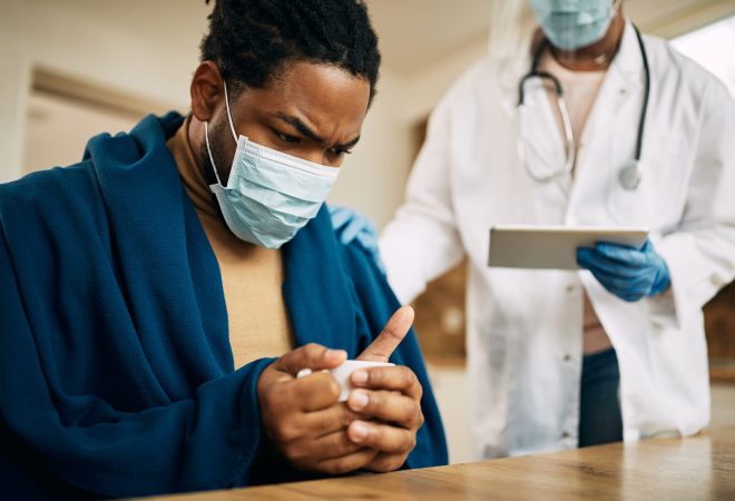 Worried black man with face mask sitting wrapped in blanket while doctor is visiting him at home.