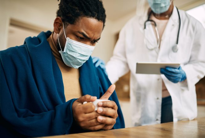 Worried black man with face mask sitting wrapped in blanket while doctor is visiting him at home.