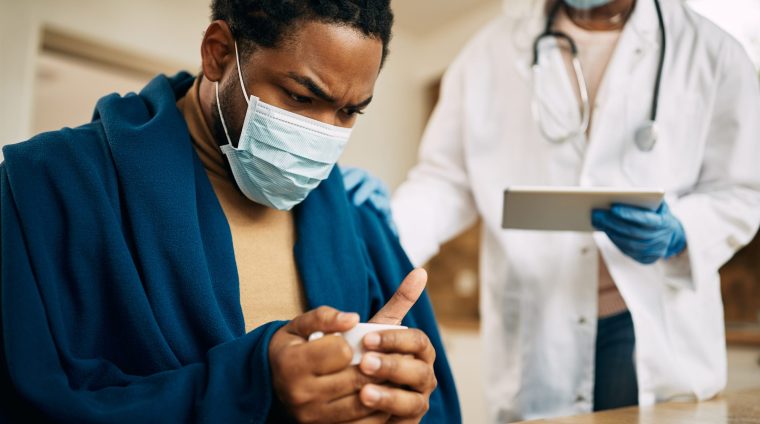 Worried black man with face mask sitting wrapped in blanket while doctor is visiting him at home.